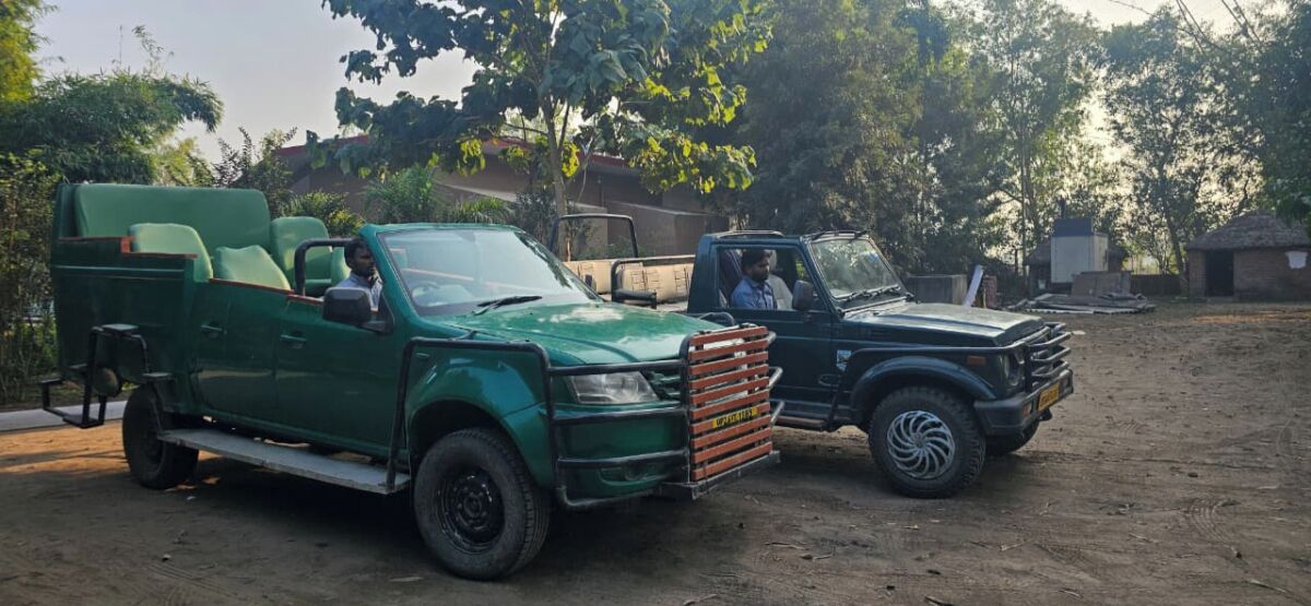 Two pickup trucks parked on a dirt surface with trees and buildings in the background.