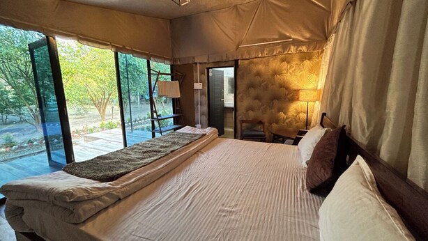 A tent-style bedroom with a bed, pillows, and a bedside lamp. There is a window with a view of trees and a door leading outside. - Earth Home Pilibhit (Puranpur)