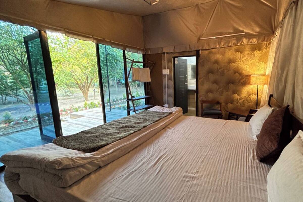 A tent-style bedroom with a bed, pillows, and a bedside lamp. There is a window with a view of trees and a door leading outside.