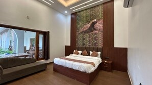 A bedroom with a large bed, a sofa, a wooden headboard, a nightstand, and a large window. - Earth Home Pilibhit (Puranpur)