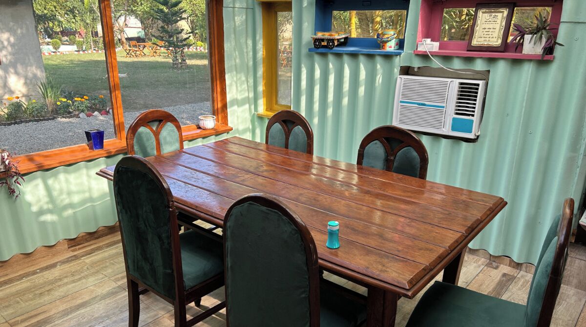 A dining area with a wooden table and chairs, a window with a view of a garden, and an air conditioning unit on the wall.