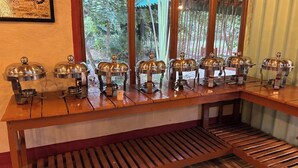 A buffet setup with multiple food service stations, each with a metal serving tray and a dome cover, arranged on a wooden table. - Earth Home Pilibhit (Puranpur)