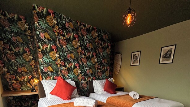 En dubbelsäng med vita sängkläder och orangea plädar, en sänggavel med ett blommönster och en hängande lampa.