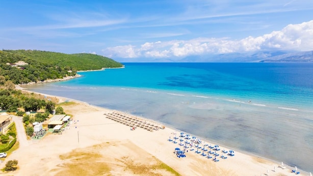 A beach with clear turquoise waters, a sandy shore, and a row of blue umbrellas. - Ormos Beach House in Apraos (Corfu)