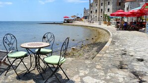 A coastal area with stone tables and chairs, a stone building, and red umbrellas. - Holiday Home With Garden and BBQ in Umag (Umag)