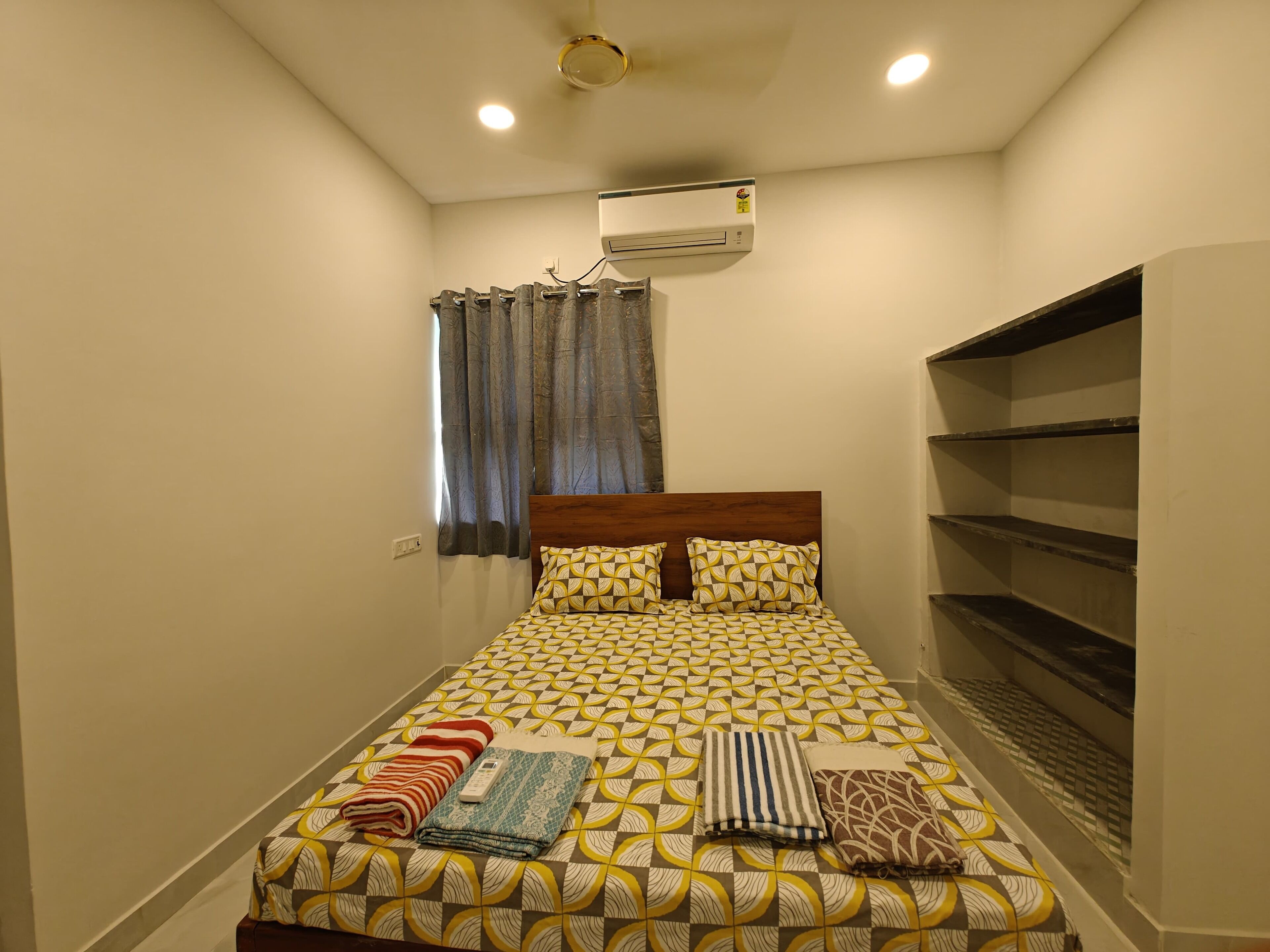 A small, neatly made bed with patterned bedding, a window with curtains, an air conditioning unit, and a built-in shelf.