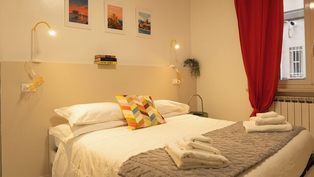 Ein ordentlich bezogenes Bett mit weißen Leinen, einem farbenfrohen Kopfkissen und einem gefalteten Handtuch auf einer grauen Decke. Zwei Wandleuchten, ein roter Vorhang und ein Fenster mit Blick auf ein Gebäude sind zu sehen.