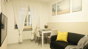 Ein Zimmer mit Esstisch, einer schwarzen Couch mit gelbem Kissen, einer grauen Decke und einem Fernseher an der Wand.