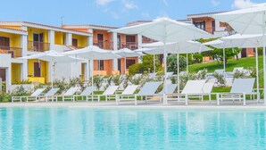A poolside area with white lounge chairs and umbrellas, a clear blue pool, and buildings with orange and yellow facades in the background. - A sophisticated space designed to accommodate up to four people, where comfort and style meet. The living room features a comfortable double sofa bed, while the bedroom, equipped with a double bed, offers an intimate and relaxing atmosphere. The modern b (Costa Serena)