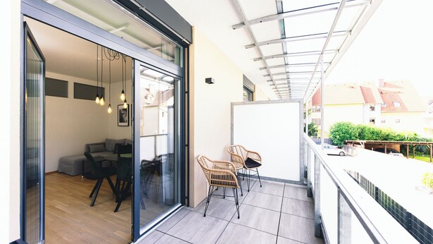 A balcony with a glass door leading to a room with a sofa and chairs, a car parked outside, and a view of buildings and greenery. - GH Luxury Apartments Graz, Kalvarienberg (Graz)