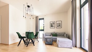 A modern living room with a dining area, a sofa, and a bed. There are two framed pictures on the wall, a chandelier, and a window with curtains. - GH Luxury Apartments Graz, Kalvarienberg (Graz)