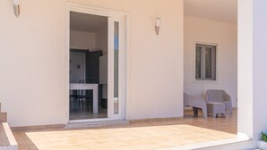Eine offene Tür führt in einen Essbereich mit Stühlen und einem Tisch, eine kleine Sitzgelegenheit im Freien ebenfalls mit Stühlen, und ein Fenster an der rechten Wand.