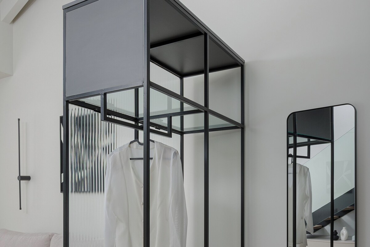 A modern, minimalist room with a built-in wardrobe featuring multiple shelves and hanging space, a small seating area with a bench, and a large mirror on the wall.