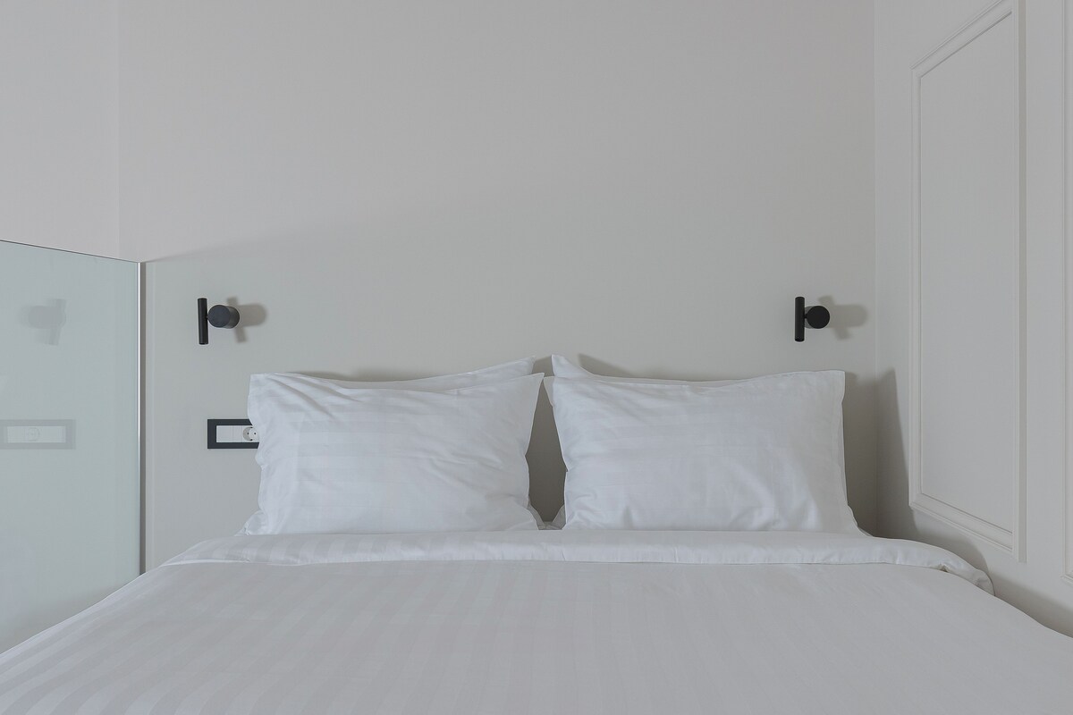 A minimalist hotel room with a large bed, white bedding, and a glass partition wall.