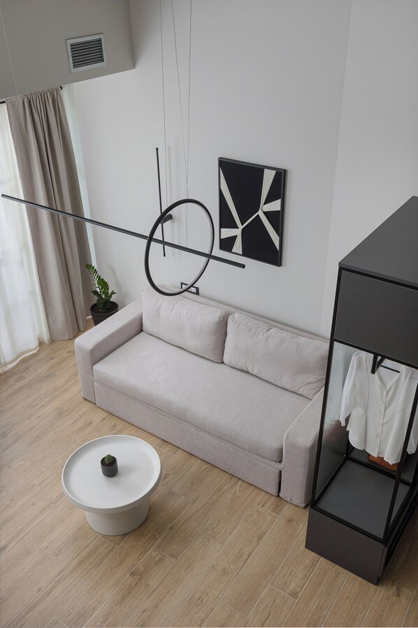 A modern living room with a light-colored sofa, a round coffee table with a plant, a black metal shelf with clothes, and a minimalist wall art piece. - OPAL Lake Suites (Ioannina)