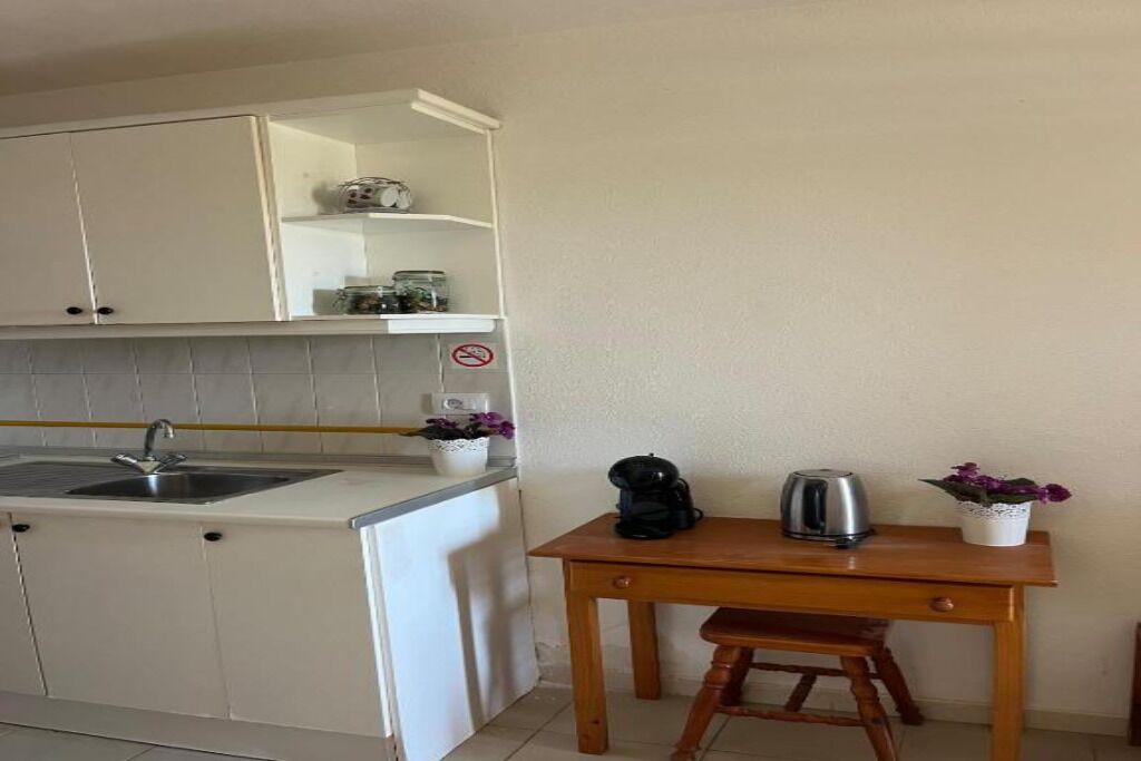 A compact kitchen with a sink, cabinets, and a small table with a kettle and flowers.