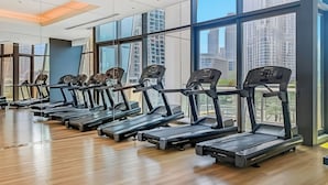 Un gimnasio con varias cintas de correr alineadas frente a grandes ventanales, con vistas a edificios altos.