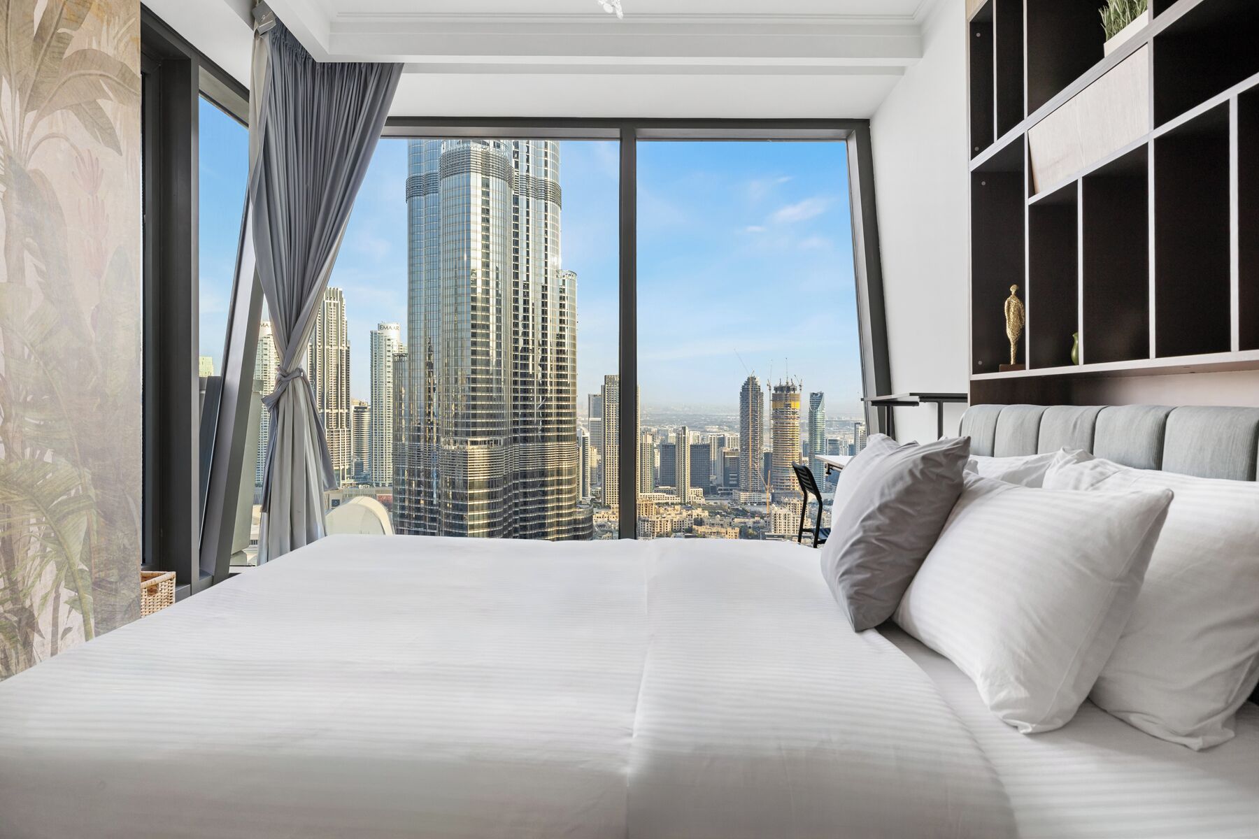 A modern hotel room with a large bed, a view of a city skyline, and a large window with curtains.