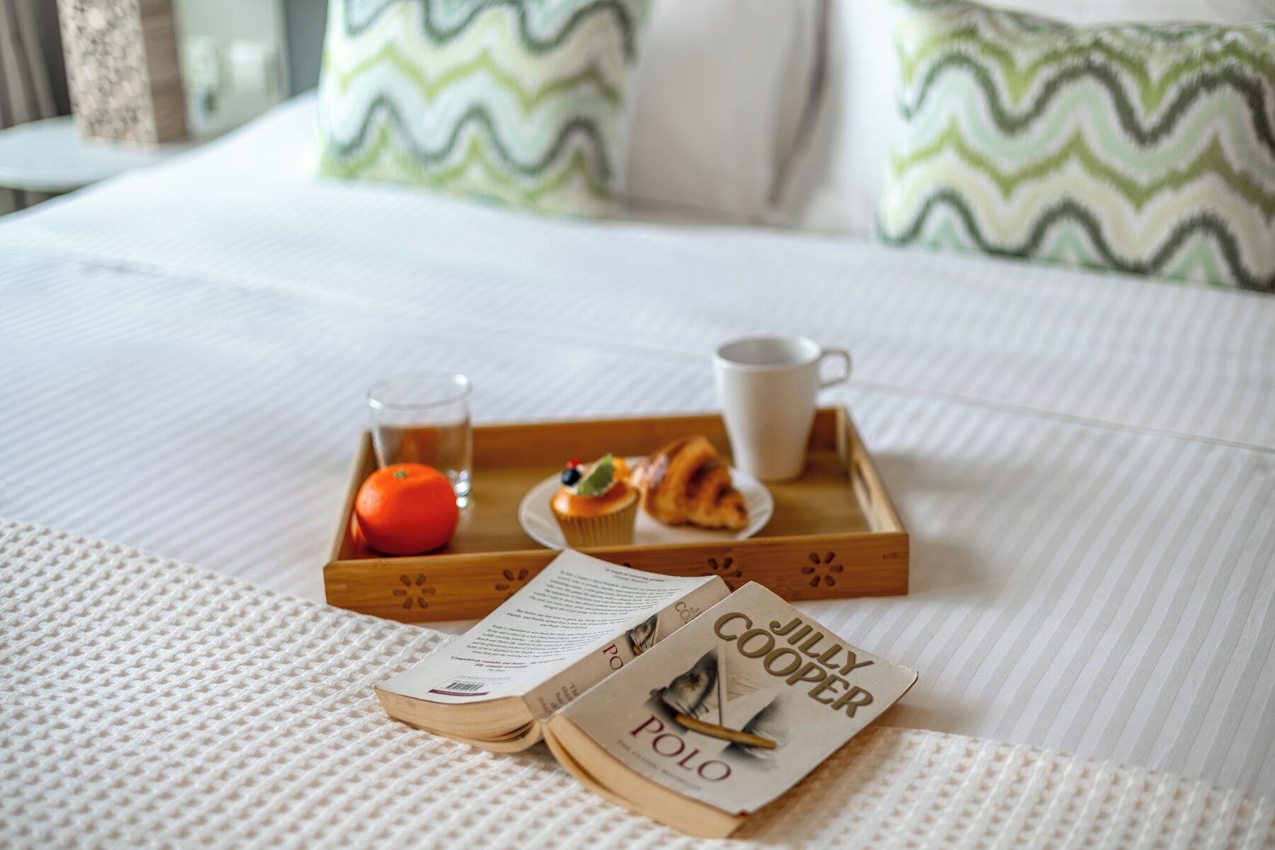 A neatly made bed with a white textured bedspread, a tray with breakfast items, a book titled 'Polo' by Jilly Cooper, and two pillows with a green zigzag pattern.