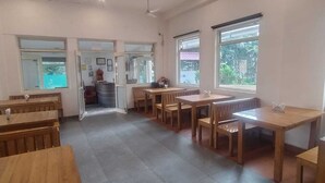 A spacious room with multiple wooden tables and benches, large windows providing natural light, and a visible staircase leading to another area. - Hotel Aviral Ganga (Rishikesh)