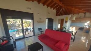 A spacious living area with a red sofa, a black coffee table, and a dining table with chairs. A kitchen is visible in the background, and there is a sliding glass door leading to an outdoor area with a pool. - Casa Quatre Brises (L'Escala)