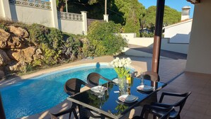 A dining table with chairs and a vase of white flowers, set by a pool with a tiled patio and a stone wall in the background. - Casa Quatre Brises (L'Escala)