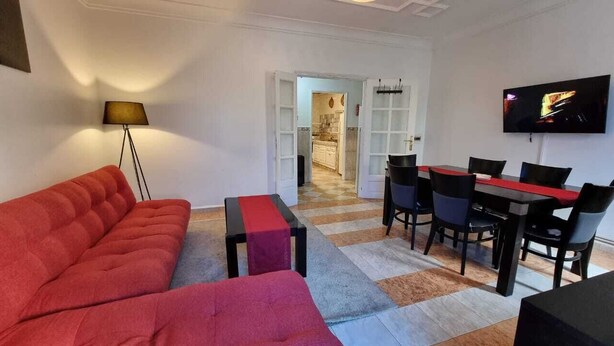 Sala de estar com sofá vermelho, mesa de centro e televisão na parede. Área de jantar com mesa e cadeiras, e cozinha visível através de uma porta aberta.