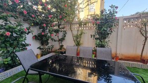 Mesa com tampo de vidro e quatro cadeiras, rodeada por um jardim com plantas em vasos e uma parede com detalhes decorativos.