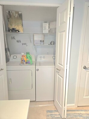 A laundry room with two washing machines, a shelf with a laundry detergent box, and a door leading to another room. - 5 HOSPITALS WITHIN 1O MILE RADIOUS Luxurious 1Bed EXTRA PLUSH! NO PET FEES! (Columbus)