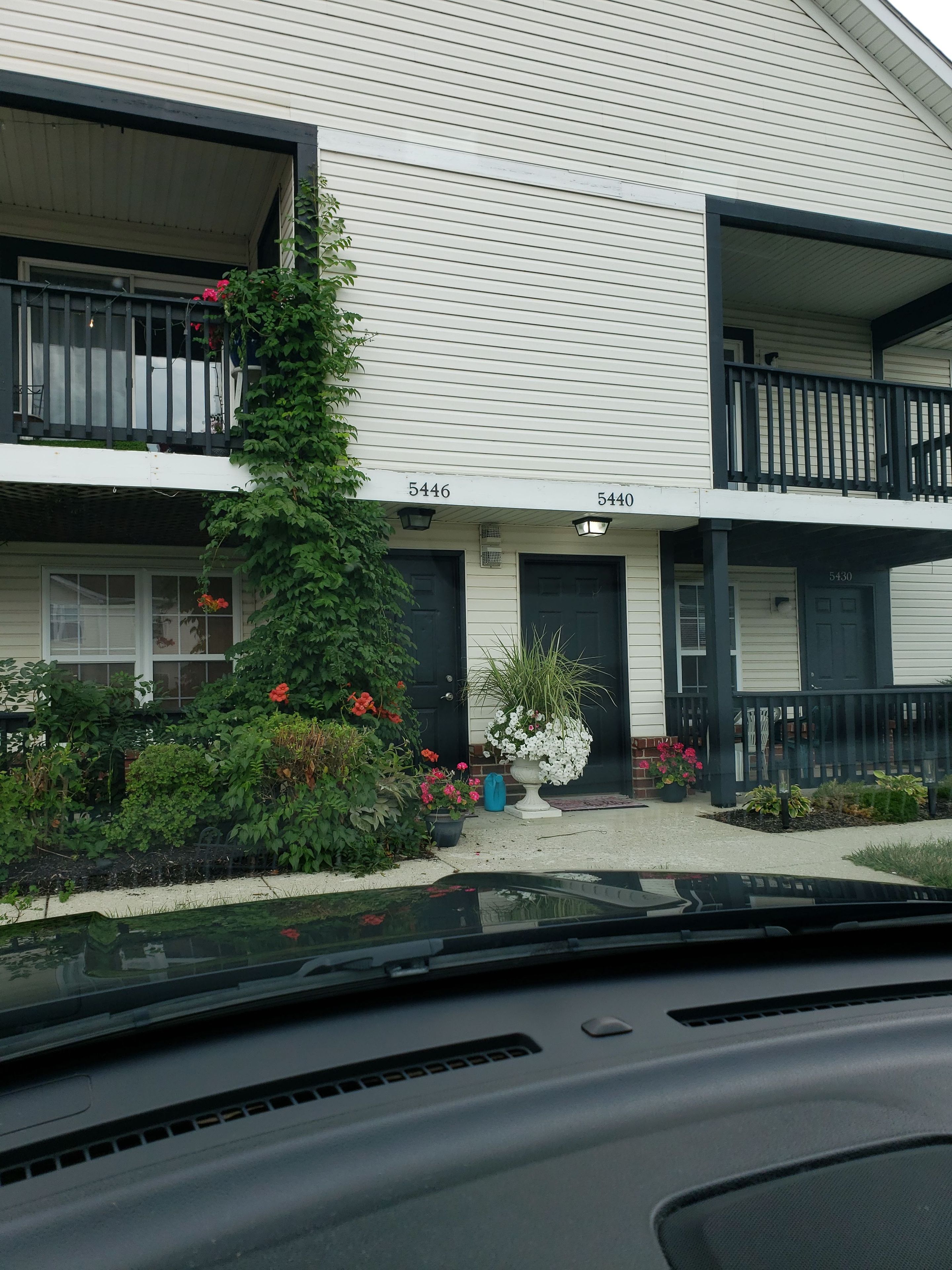 A two-story building with numbered doors, a small garden with various plants and flowers, and a visible balcony.