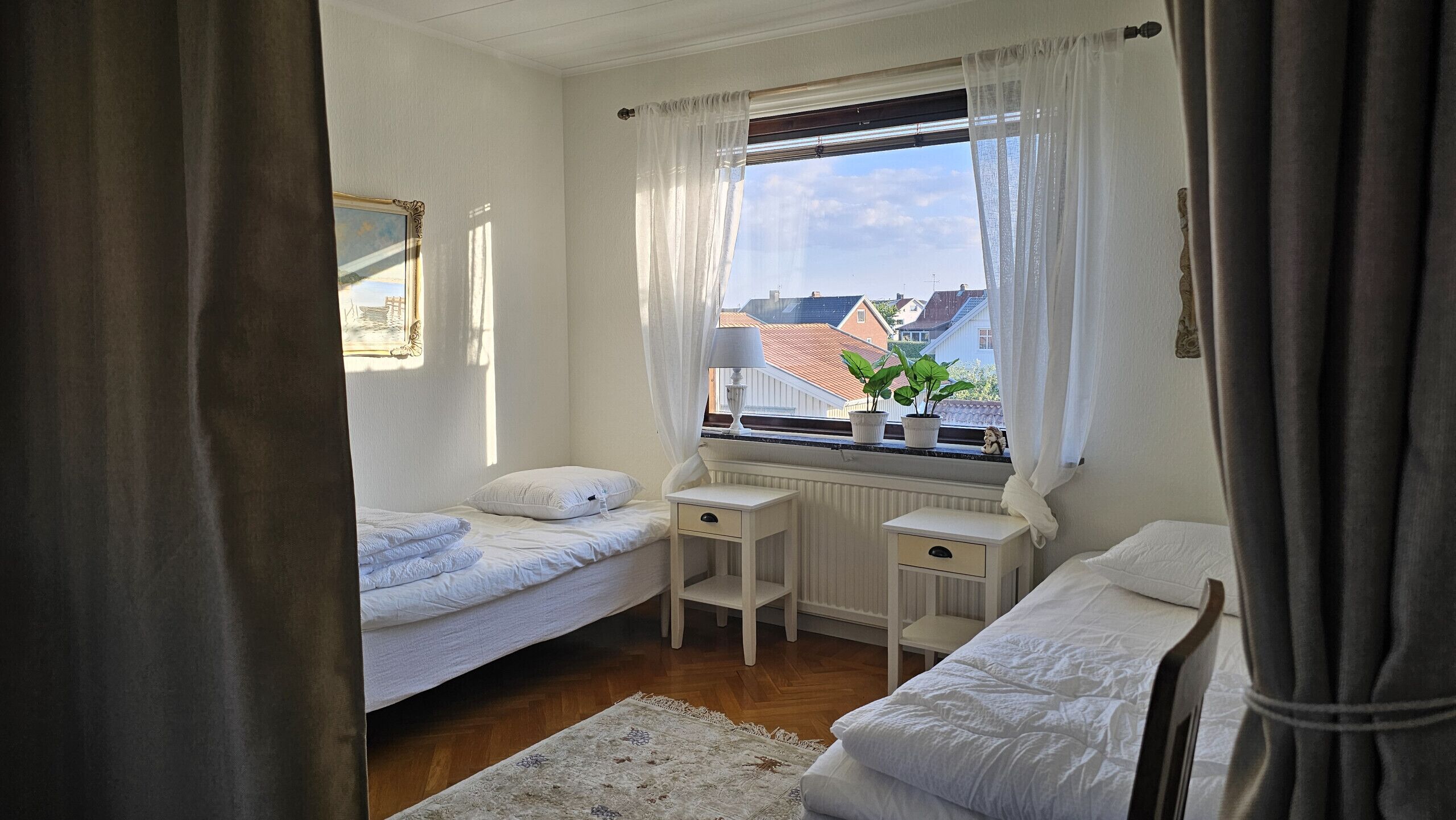 A small, tidy bedroom with two single beds, a window with curtains, a radiator, and a small table with a lamp.