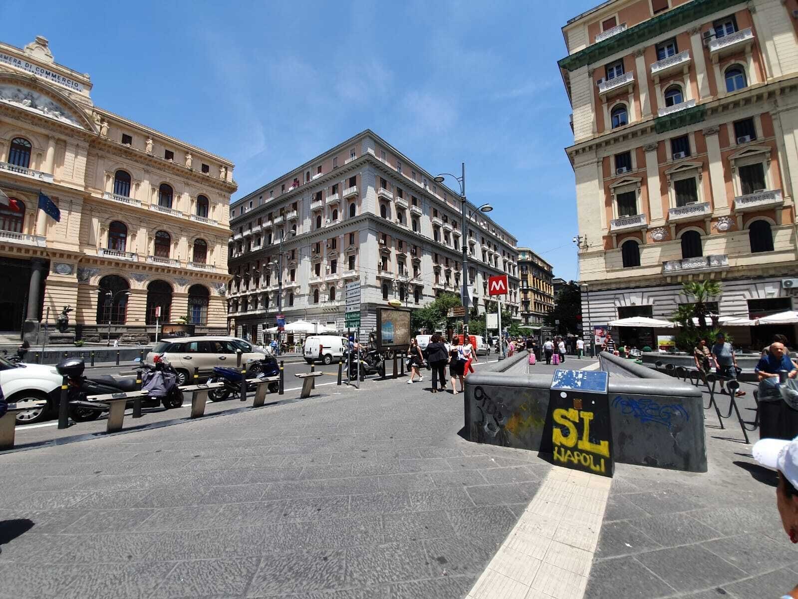 A busy urban square with historic buildings, a central monument, and a clear sky.