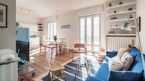 A modern living room with a blue sofa, a glass coffee table, and a dining area with a white table and chairs. - Infinito (Como)