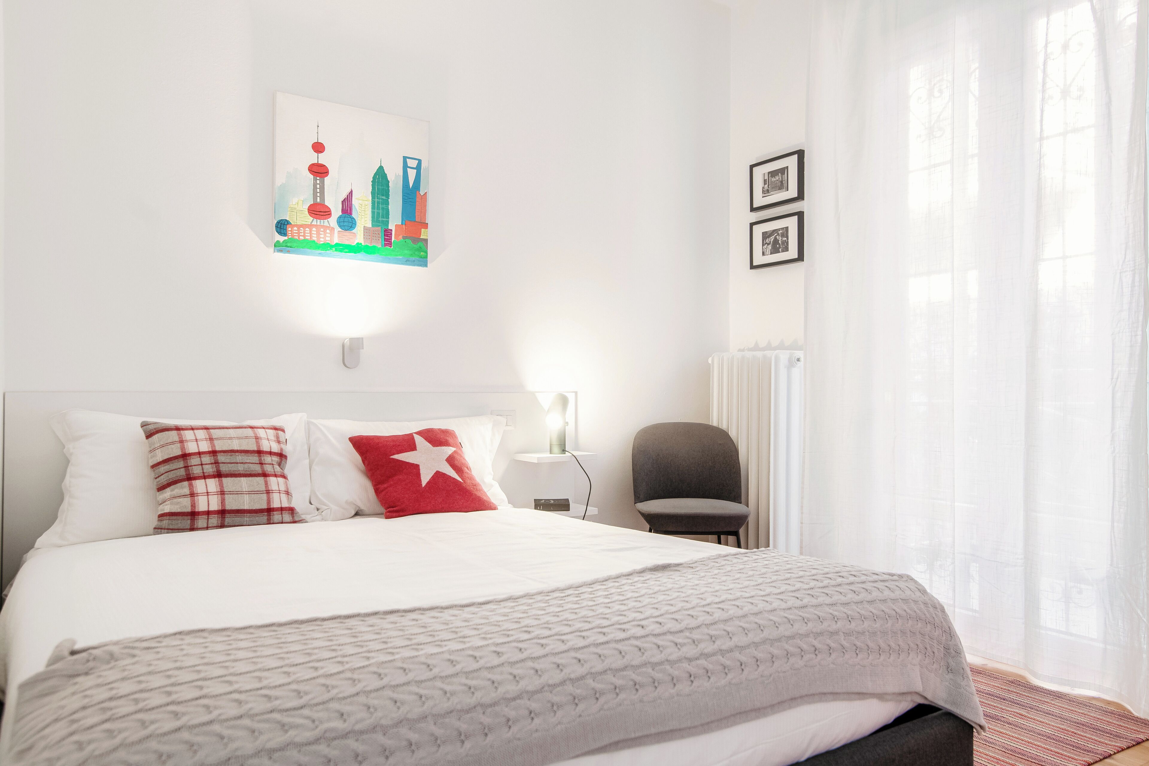 A hotel room with a bed, a chair, a wall art piece, and a window with sheer curtains.