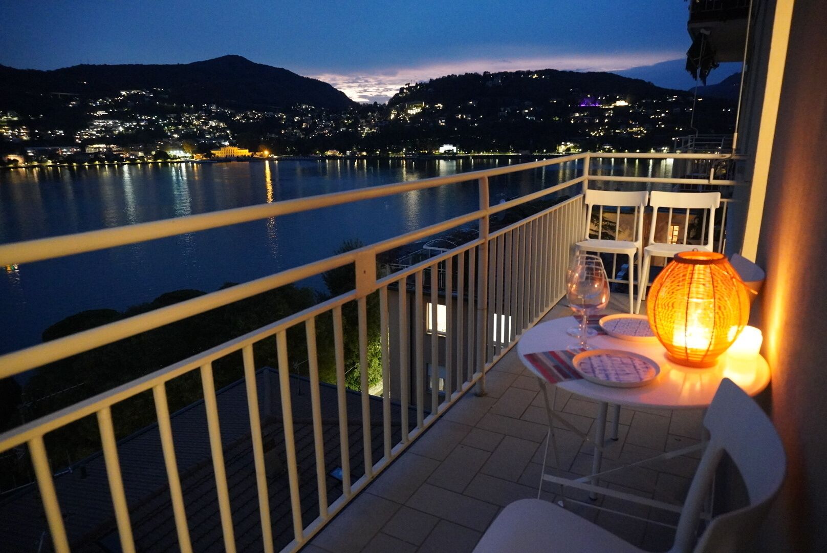 A balcony with a table set for two, overlooking a body of water and a mountainous landscape at dusk.