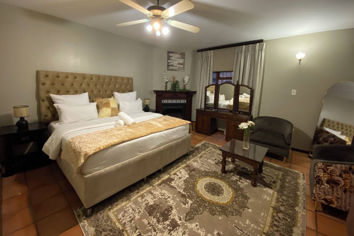 A hotel room with a large bed, a ceiling fan, a dresser, a mirror, a chair, and a small table with a vase.