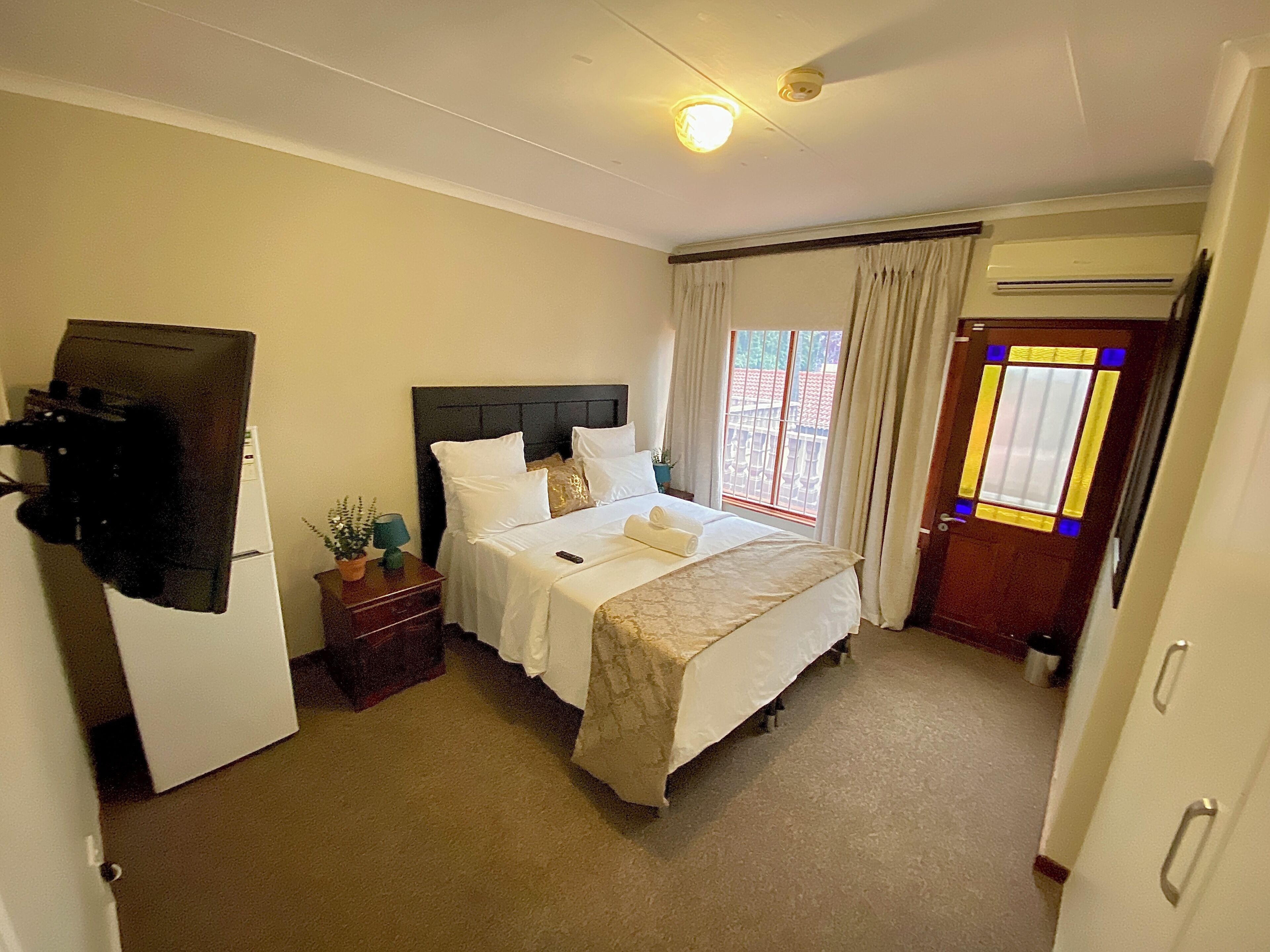 Habitación de hotel con cama, mesita de noche, lámpara, planta en maceta y puerta con ventana.