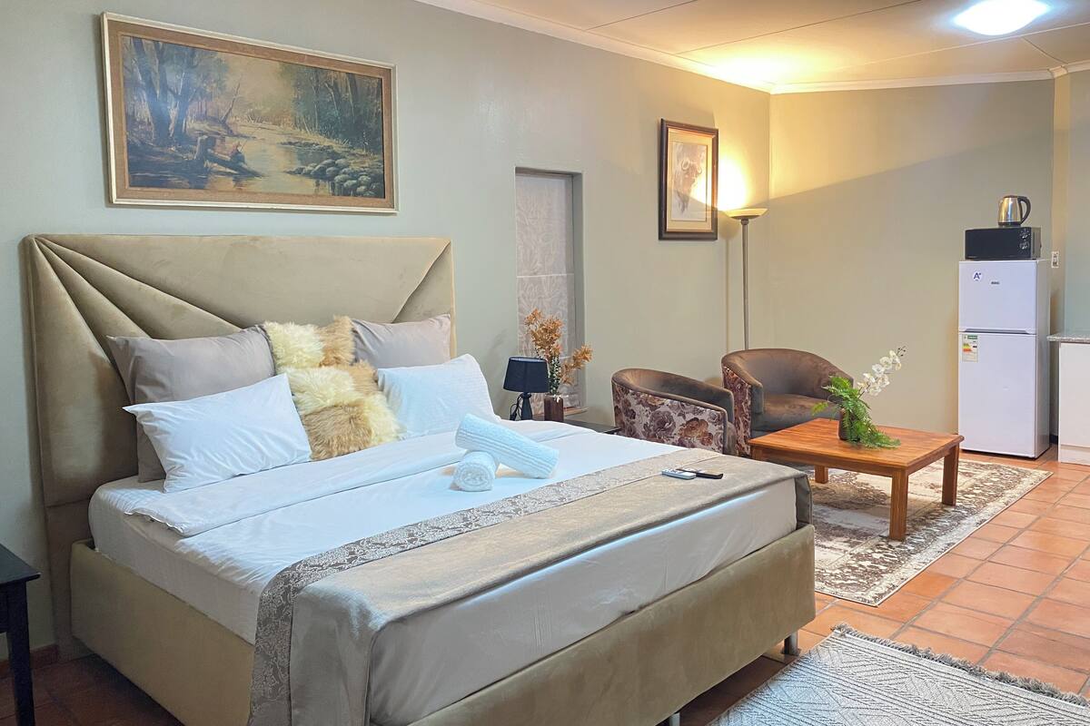 A neatly arranged bedroom with a large bed, a bedside table, a chair, a small table with a vase, a painting on the wall, and a refrigerator.
