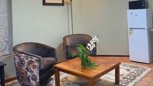 Una habitación con un sillón de cuero marrón, una mesa de madera con un cojín floral, un cuadro en la pared, una lámpara de pie y un refrigerador blanco con una tetera encima.