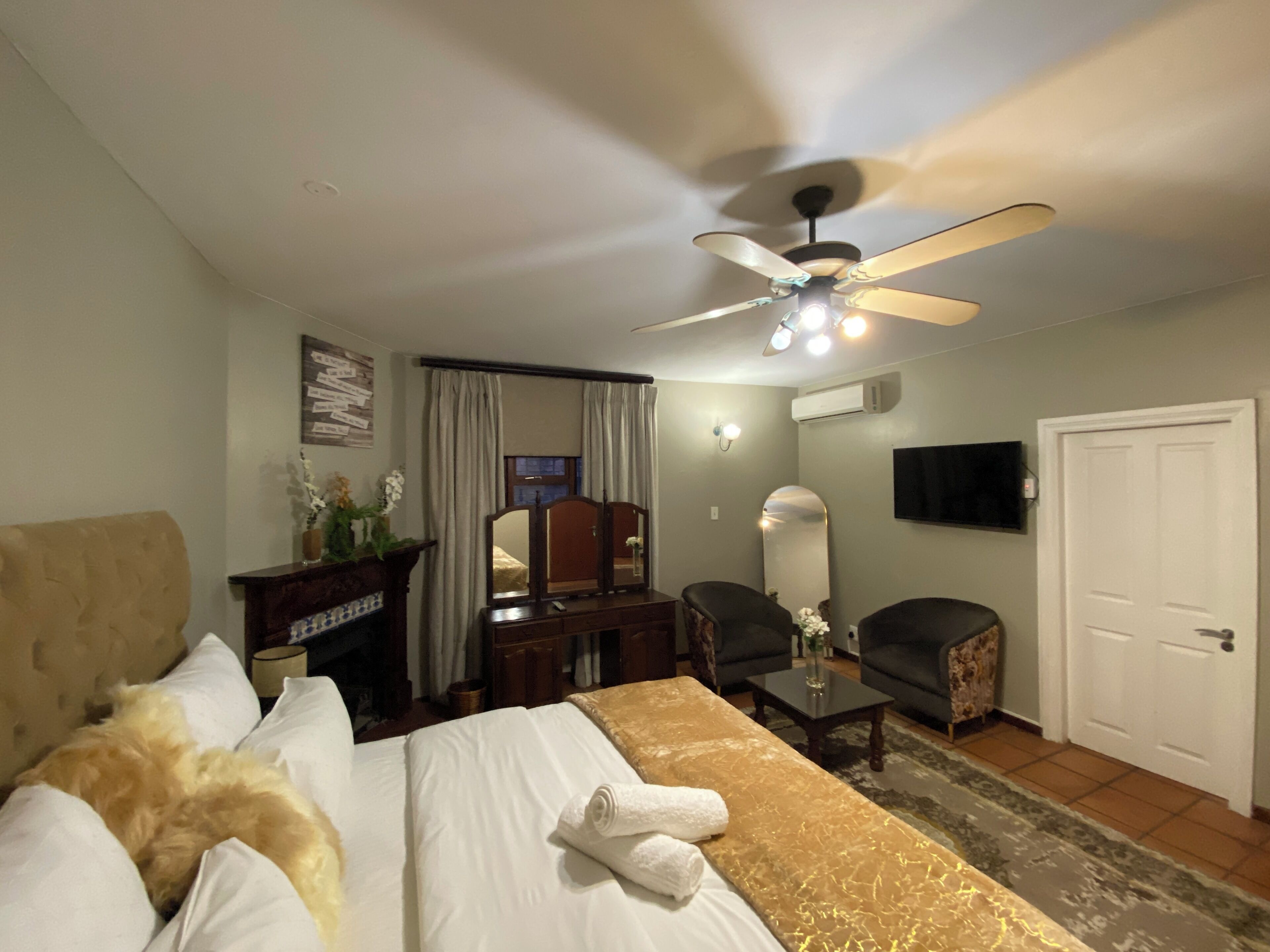 A hotel room with a bed, a desk, a chair, a ceiling fan, an air conditioning unit, a television, and a door.