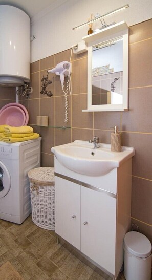 Une salle de bain avec un lavabo blanc, un miroir et un sèche-cheveux fixé au mur. On y trouve également un lave-linge et un panier en osier posé sur le sol.