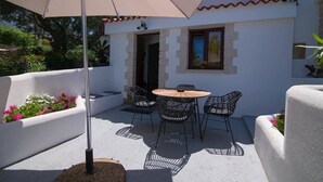 Une terrasse avec une table ronde et quatre chaises, un parasol et un mur blanc avec une fenêtre.