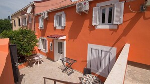 A two-story building with orange walls, white shutters, and tiled roofs. There are satellite dishes, an air conditioning unit, and a small outdoor seating area with a table and chairs. - Luisa Rooms / Luisa room B2 (Veli Lošinj)