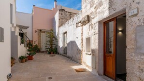 A narrow stone-paved courtyard with a wooden door, potted plants, and a small staircase leading to a pink building. - Dimora Giulietta, modern accommodation with vaulted ceilings, in a Salento courtyard (Miggiano)