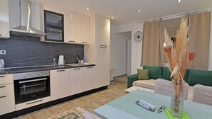Et moderne køkken med hvide skabe, en sort bordplade og en indbygget ovn. Der er et spiseområde med et bord og stole, og en grøn sofa i baggrunden.