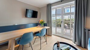A modern dining area with a wooden table, blue chairs, a flat-screen TV, and a large window with curtains. - Dependance Windrose 174 (Wenningstedt-Braderup (Sylt))