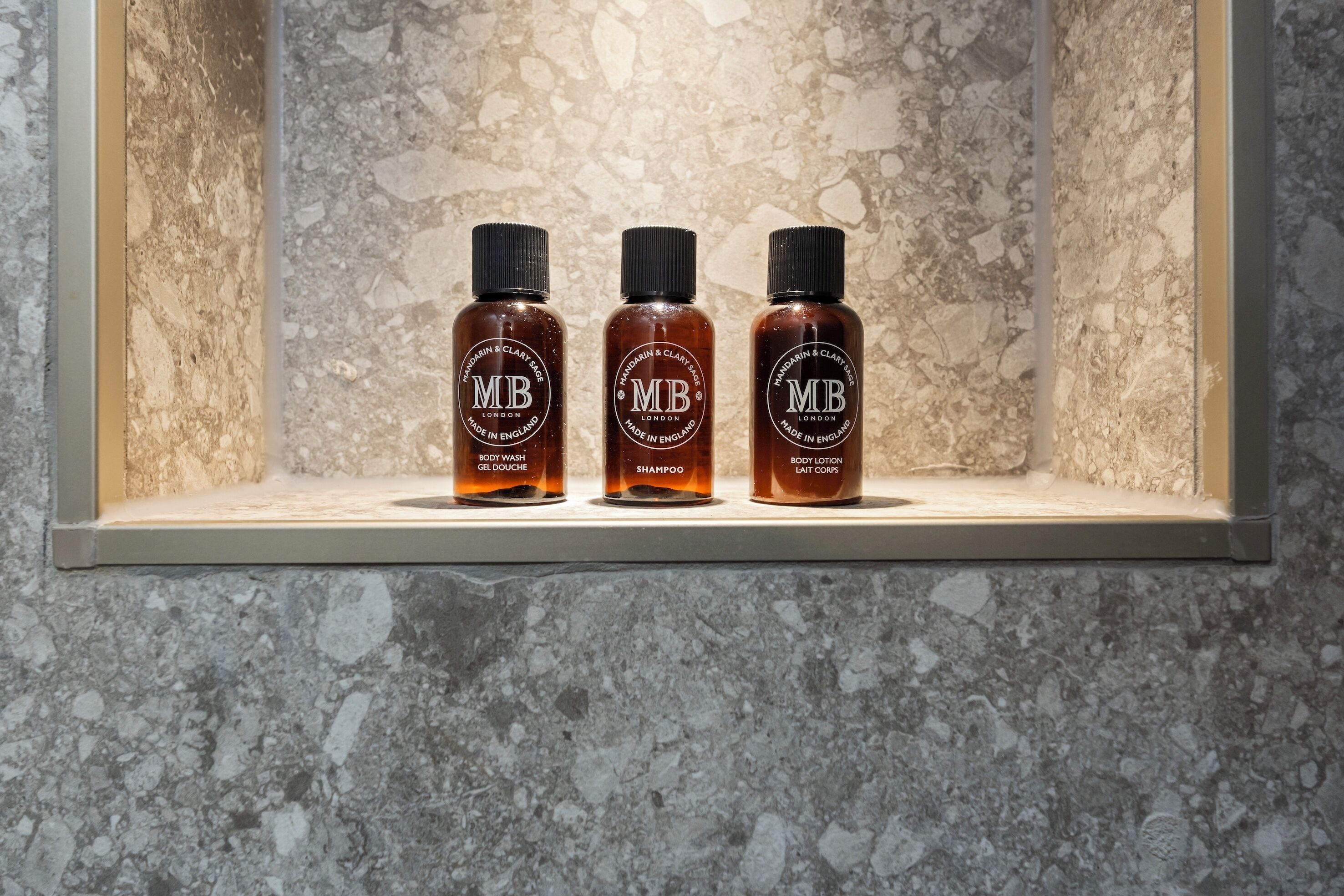Drei Flaschen Duschgel, Shampoo und Körperwaschmittel auf einem Regal vor einer strukturierten Wand.