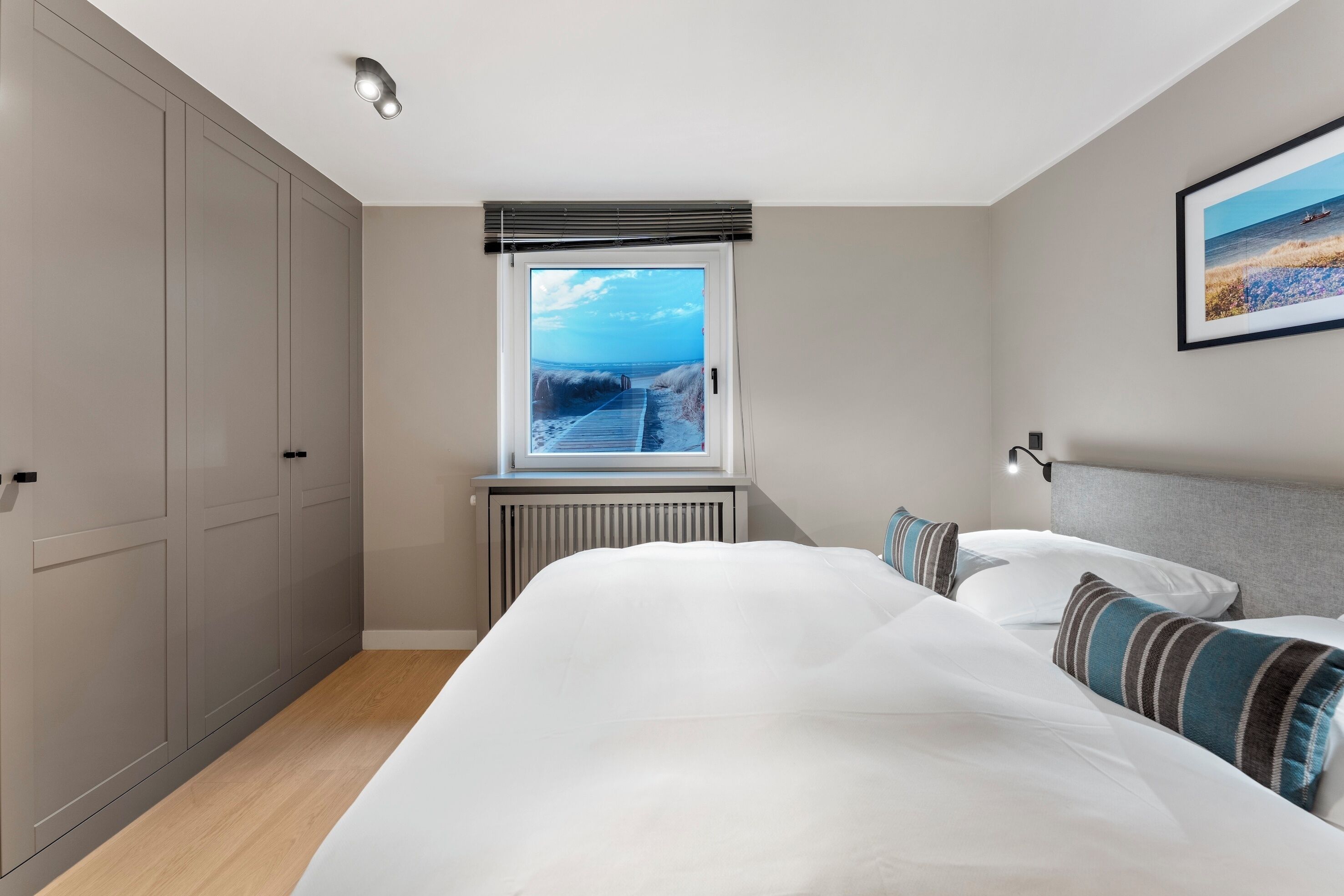 Ein Schlafzimmer mit einem großen Bett, Blick auf den Strand durch ein Fenster und einem Heizkörper unter dem Fenster.