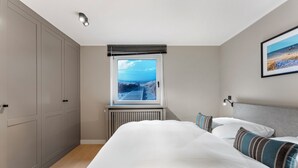 A bedroom with a large bed, a view of the beach through a window, and a radiator below the window. - Dependance Windrose 174 (Wenningstedt-Braderup (Sylt))