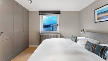 Ein Schlafzimmer mit einem großen Bett, Blick auf den Strand durch ein Fenster und einem Heizkörper unter dem Fenster.